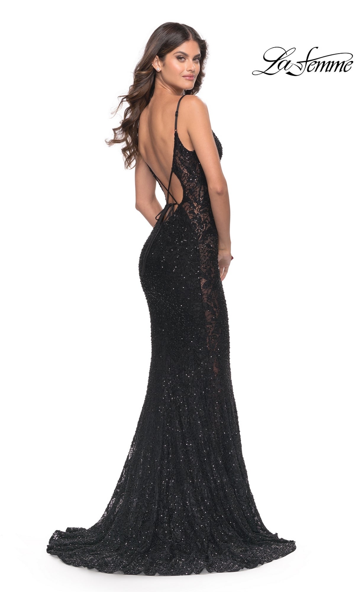 A woman in the La Femme Beaded Long Black Lace Prom Dress 31257—fitted, floor-length with sequins and a low open back—stands looking over her shoulder.